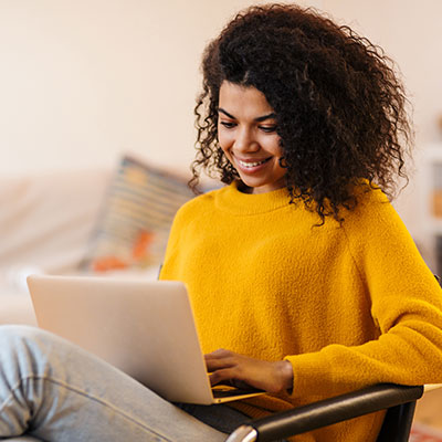 a smiling young woman with curly hair sitting on a chair looking down at a laptop in her lap